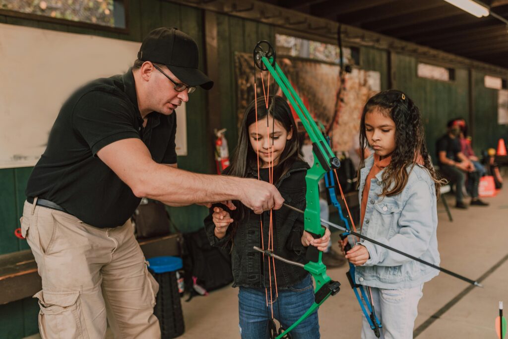 Adult male archery coach teaching adorable little multiethnic girls to shoot with bow and arrow during classes in range